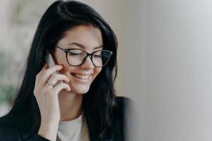 Happy woman lawyer keeps mobile phone near ear, has pleasant conversation with client, discuss working issues, smiles gently, wears transparent glasses, formal wear. Female employee calls someone photo