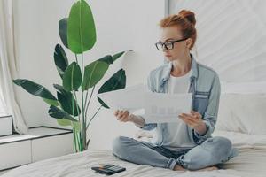 Redhead serious Caucasian woman sits in lotus pose on bed studies documents calculates bills photo