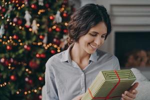 feliz mujer hispana complacida sosteniendo una caja de regalo de navidad mientras se sienta en un sofá cerca del árbol de navidad foto