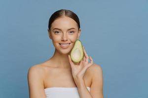 Horizontal shot of happy brunette woman holds half of avocado near face uses anti aging organic recipes stands refreshed indoor wrapped in towel isolated over blue background. Healthy diet concept photo