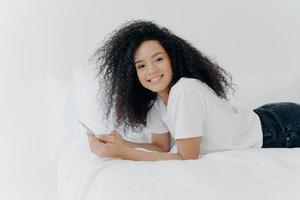 mujer bastante alegre con corte de pelo afro, disfruta chatear y hacer contactos, se acuesta en una cama cómoda, mira fotos, tiene una expresión facial relajada, aislada sobre fondo blanco. comunicación en línea foto