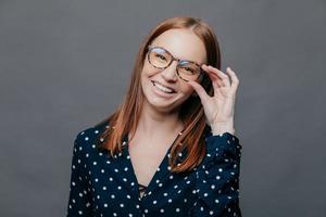 Smiling woman with pleased facial expression, keeps hand on rim of spectacles, wears black polka dot blouse, rejoices promotion at work, poses over grey background. People, happiness concept photo