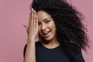 Happy teenage girl has fun and hides face with palm, has joyful expression, curly bushy hair, dressed in casual black clothes, isolated on pink background. Shy glad young woman gets compliment photo