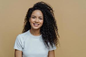 Horizontal shot of curly haired woman with healthy skin, toothy smile, enjoys being photographing, wears casual white t shirt, isolated on beige background. People, emotions and lifestyle concept photo
