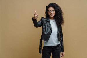 Confident cheerful African American woman shows thumb up in approval, gives recommendation or advice dressed in stylish leather jacket smiles positively isolated on beige background. I like and accept photo
