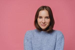 Beautiful brunette lady in knitted warm sweater looks directly at camera, smiles positively, enjoys nice winter day, isolated over pink background, has pleasant conversation with interlocutor photo