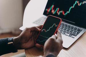 Male hands holding smartphone next to computer screen analyzing financial stock market charts photo