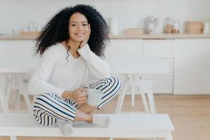 Indoor shot of pretty African American woman wears white jumper, striped pants, socks, poses on bench with cup of tea spends leisure at home in cozy kitchen feels relaxed. Female enjoys morning coffee