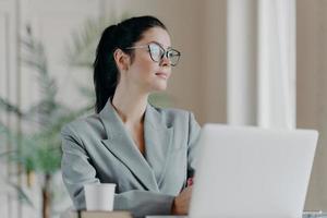 Successful brunette female entrepreneur going to watch webinar via laptop computer, concentrated aside with thoughtful expression, wears transparent glasses, dressed in formal clothes, drinks coffee photo