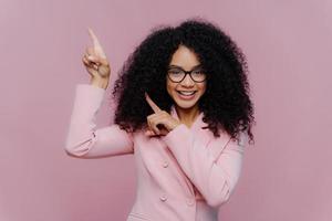 Half length shot of cheerful Afro American woman with dark bushy hairstyle, wears violet suit, points above with both index fingers, demonstrates way to something, invites you going upstairs