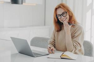 una mujer de jengibre positiva con ropa informal graba un mensaje de audio o un teléfono inteligente mientras trabaja en una computadora portátil foto