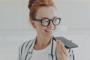 Portrait of happy cheerful red-haired woman in eyewear holding mobile phone, recording audio message photo