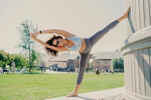 Horizontal shot of flexible fit woman stands on one leg leans with arms raised stays in good physical shape does gymnastics exercises poses outdoor enjoys good weather. People wellness concept photo