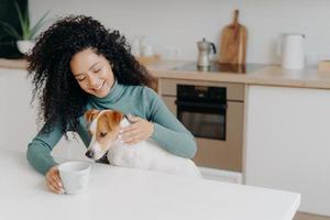 Happy Afro African woman with curly hairstyle treats dog in kitchen, pose at white table with mug of drink, enjoy domestic atmosphere, have breakfast together. People, animals, home concept. photo