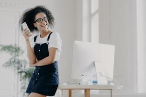 Happy curly haired girl uses mobile phone for communication and keeping in touch, wears spectacles and casual clothes, leans at desktop, poses in spacious cabinet. Glad student uses technology photo