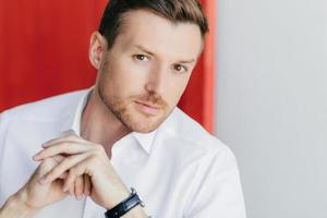 Cropped shot of serious young male CEO or company owner, has success, climbs career ladder, looks with strict expression at camera, keeps hands together, poses against red and white background photo