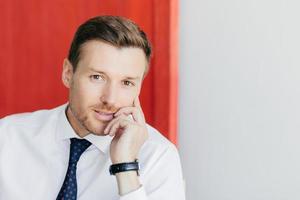 Photo of attractive young businessman with confident expression holds chin, dressed in formal white shirt, has watch on arm, thinks about starting new business, poses against red and white wall