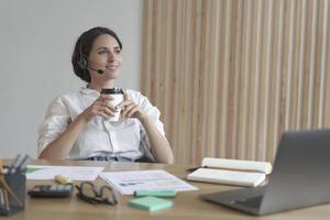 Pleasant italian business woman in headset sits at desk at home office with takeaway coffee photo