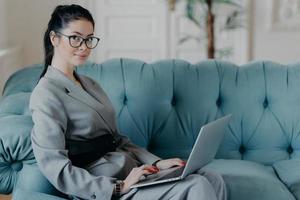 Successful female financer updates data in application on modern laptop computer, looks seriously at camera, sits on comfortable sofa, owns project of web store, wears formal outfit optical spectacles photo