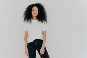 Photo of smiling woman with curly dark hair, has slim figure, wears white t shirt and black jeans, has glad face expression, models against white background, copy space for your advertising content