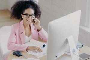 mujer positiva de piel oscura con cabello rizado y peinado, teclados en la computadora, involucrada en el proceso de trabajo, usa anteojos y ropa elegante, tiene conversación telefónica, posa en el lugar de trabajo.