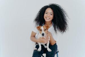Photo of glad dark skinned female being forever together with pedigree puppy, enjoys leisure at home, sits on chair, isolated over white background. Human and animal connection. Trust and friendship