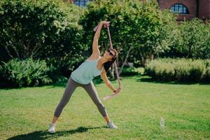 una foto completa de una mujer activa estira los brazos con goma de mascar tiene buena flexibilidad usa polainas de camiseta y zapatillas de deporte tiene entrenamiento con poses de banda de resistencia al aire libre en el césped verde durante el verano