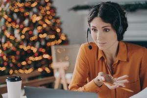 Young businesswoman in headset talking with colleagues during online meeting in office at christmas photo