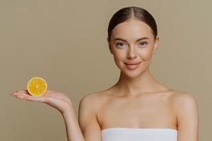 Portrait of lovely dark haired woman holds slice of orange on palm prefers natural beauty products undergoes spa treatment has skin care routine wrapped in bath towel isolated over brown background photo