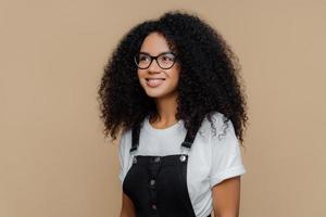 Pleased dreamy woman with curly hairstyle, looks through transparent glasses, dressed in casual white t shirt and black dungarees, poses against brown background, has good mood. People and emotions photo