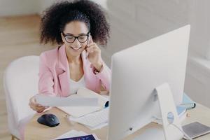 Pleased beautiful gorgeous curly haired businesswoman looks through documents, has telephone talk, sits at worplace, uses computer for making financial report, wears spectacles for vision correction photo