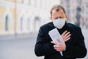 Infected man wears medical mask, has constant coughing, symptoms of Covid-19, holds rolled up newspaper, poses at city outdoor, needs isolation to stop spreading Coronavirus. Preventive measures photo