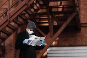 Concentrated man wears sunglasses and coat, medical mask, protective rubber gloves, reads fresh news about coronavirus spreading from newspaper, poses against blurred background with stairs. photo