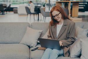 Woman in glasses sitting on cozy couch in office lounge while chatting on computer laptop photo