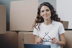 una chica hispana está sentada en el suelo con una laptop en un apartamento nuevo entre cajas de cartón. foto