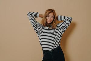 Half length shot of pretty satisfied young woman keeps hands behind head smiles gently poses indoor photo