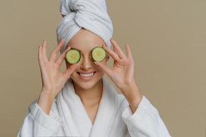 Indoor shot of pleased woman covers eyes with cucumber slices nourishes skin smiles happily dressed in bath dressing gown wrapped towel on head stands indoor. Beauty spa cosmetology concept. photo