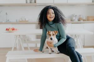 Joyful Afro woman sits at white bench together with dog against kitchen interior, table with plate full of red apples, get pleasure while playing at home. Animal owner feels care and responsibility photo
