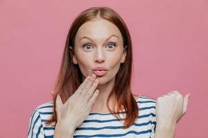 el primer plano de una mujer atractiva levanta las cejas, frunce los labios, mantiene la mano cerca de la boca, tiene el pelo castaño, sorprendido por algo, modelos sobre fondo rosa. personas, expresiones faciales y concepto de belleza. foto