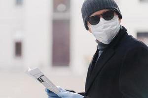 Coronavirus, health care, protection and quarantine concept. Serious man dresses hat, sunglasses, disposable medical mask and rubber gloves, concentrated somewhere, reads newspaper, poses outdoor photo