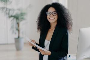 Photo of happy female entrepreneur with Afro hairstyle, studies documentation, wears spectacles and elegant clothes, stands in office interior, prepares to present her business ideas for colleagues