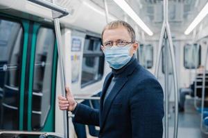 Shot of young man wears spectacles and protective face mask to prevent spreading coronavirus disease or flu epidemic in public transport, poses at empty subway carriage. Public health solution
