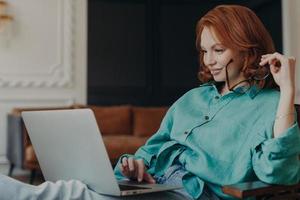 Pleased young redhead woman sits on comfortable armchair at home, does remote work, reads news in social networks, busy with computer job, concentrated in monitor of laptop. Modern technologies photo