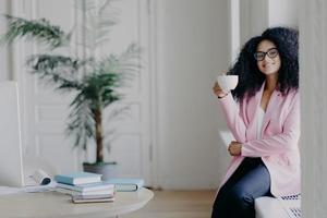 Positive African American female business worker sits at windowsill, holds mug of coffee, dressed in formal clothes, poses near desktop with books, green indoor plant in background, has break