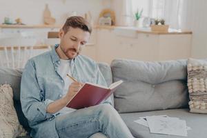 Busy young man writing in red notebook with pencil photo