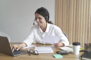 Happy woman wears headphones while sits at desk at home office looks at laptop screen works online photo