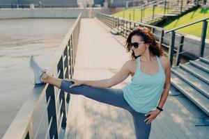 Horizontal shot of of brunette woman wears casual sport clothes stretches legs on fence has athletic body warms up before cardio training has flexible fit arms and legs poses outside. Active lifestyle photo