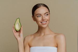 Beauty skin care concept. Positive healthy pleased woman looks tenderly aside smiles gently holds half of avocado has perfect makeup white teeth wrapped in bath towel isolated over brown studio wall photo