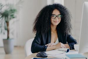 directora positiva con cabello afro rizado, reescribe información en el bloc de notas de la computadora, usa anteojos ópticos y ropa formal, bebe café, posa en el escritorio, prepara el horario de trabajo foto