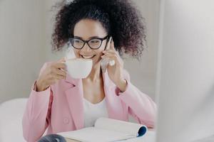 imagen recortada de una joven afroamericana feliz que hace una llamada telefónica, bebe café con leche aromático o espresso, posa en el interior de la oficina, tiene una sonrisa agradable, usa anteojos ópticos y una chaqueta formal rosada foto